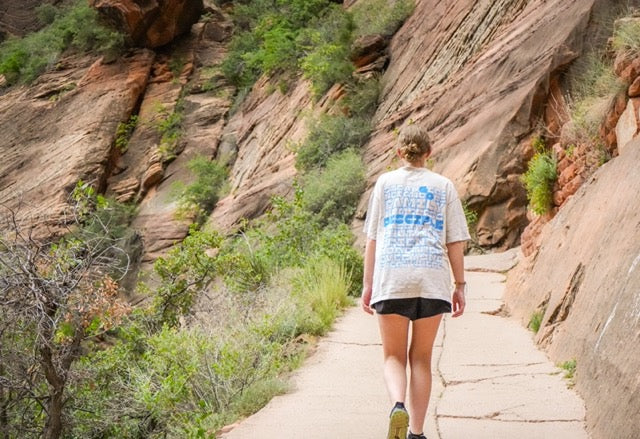 Person walking on a trail with rocky cliffs and greenery in the background