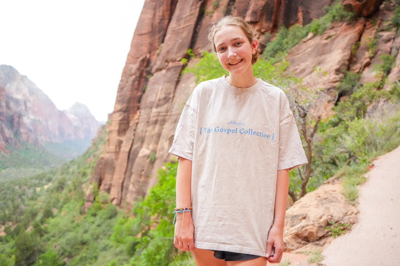 Person standing in a scenic outdoor location with mountains and greenery.