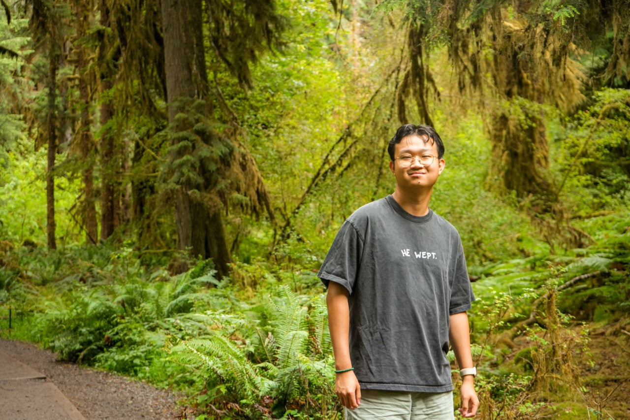 Man standing in a lush green forest wearing a gray t-shirt with text.