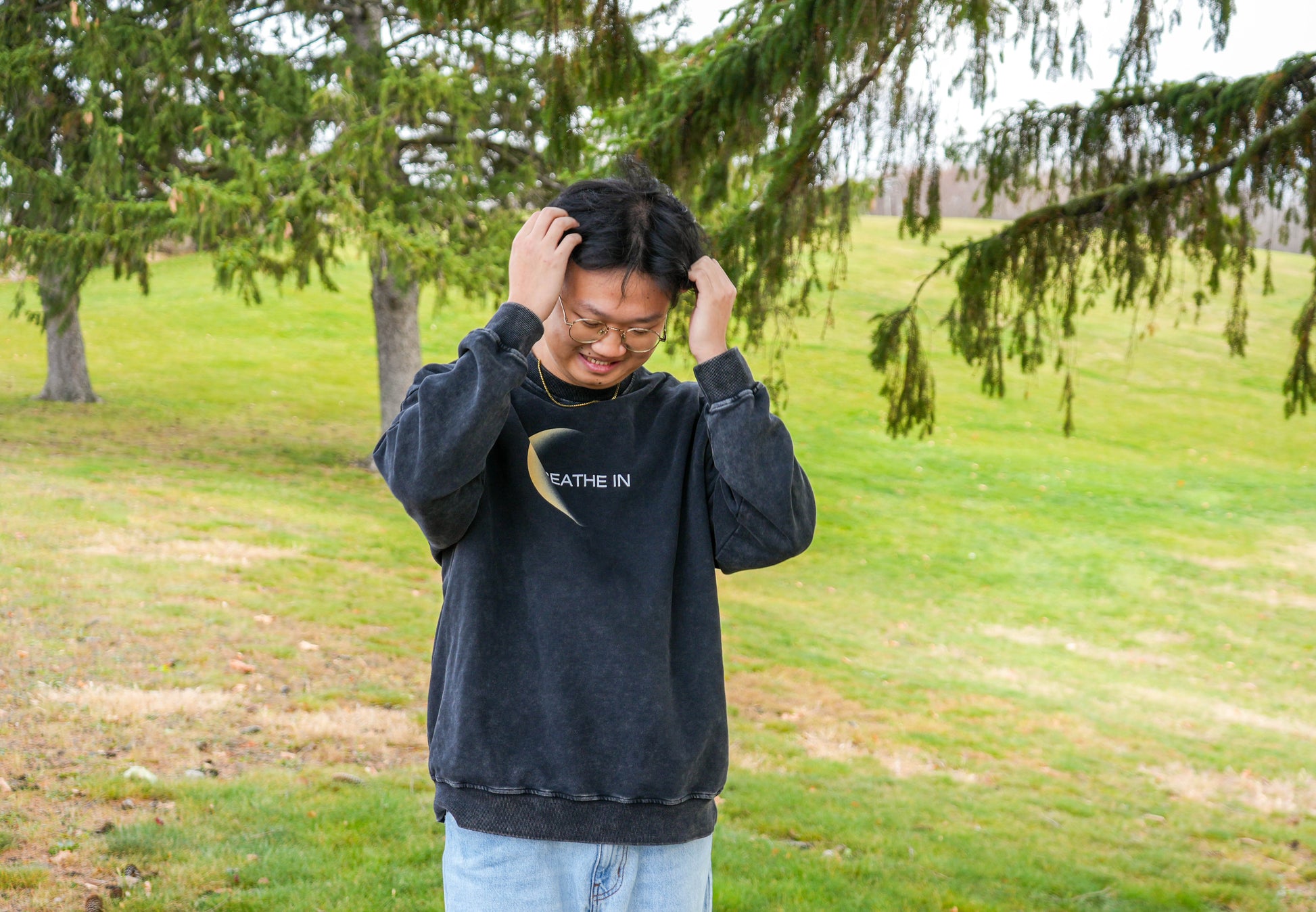 Person wearing a black hoodie with text outdoors on a grassy area with trees.