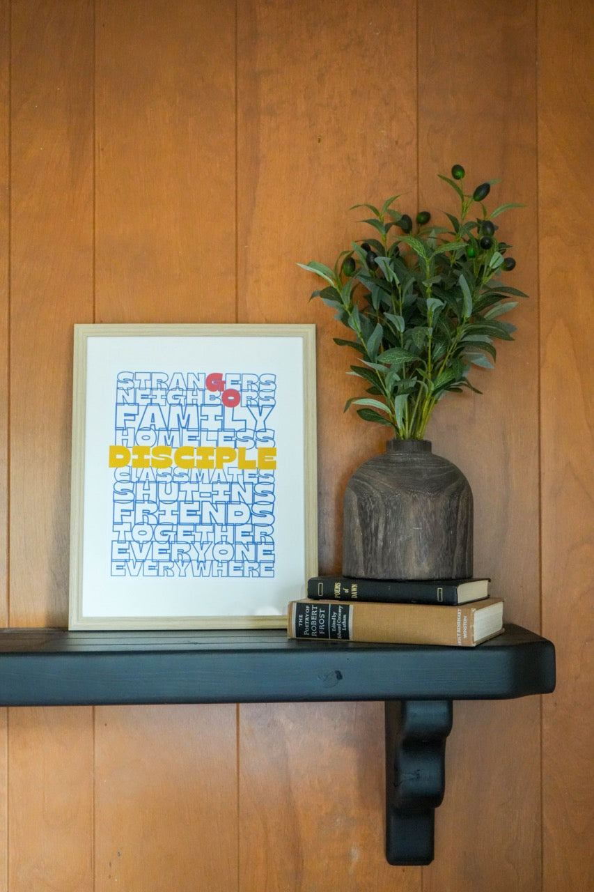 Framed artwork, plant, and books on a wooden shelf against a wooden wall.