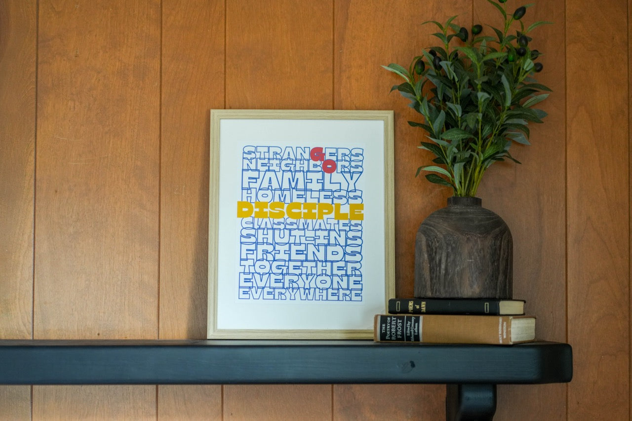 Framed artwork on a shelf against a wooden wall with a plant and books.