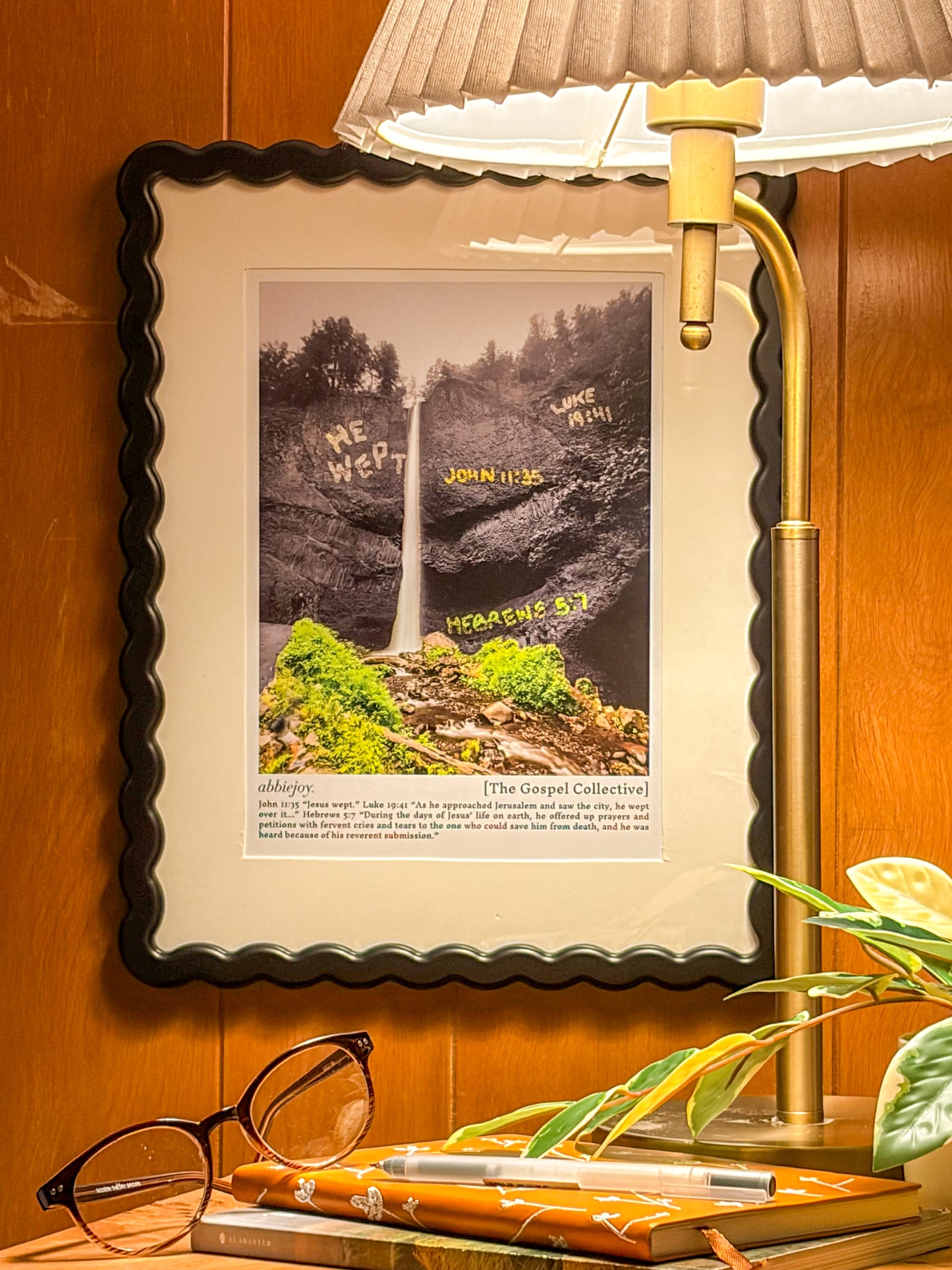Framed picture of a waterfall with text on a wooden surface next to a lamp and glasses.